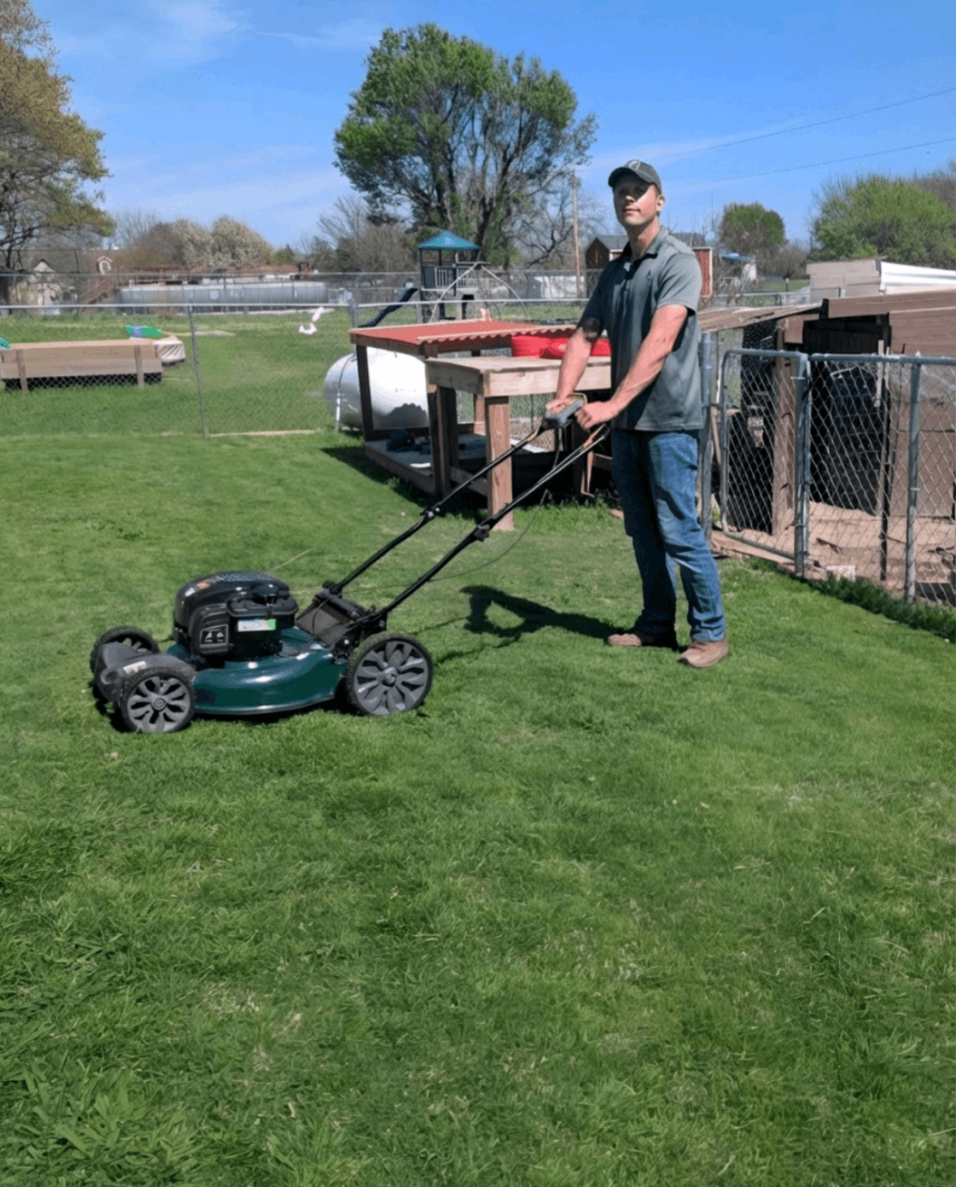 A man in a cap pushes a green lawnmower across a sunny backyard lawn.