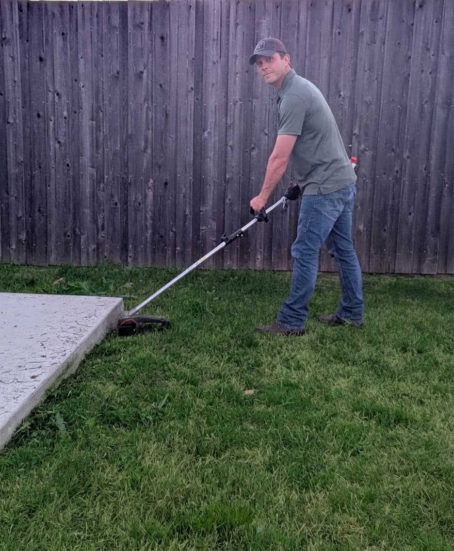Man using a string trimmer to edge a lawn next to a concrete patio.