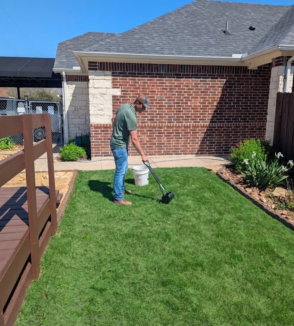 Man in a green shirt uses a rolling tool on a lush green backyard lawn.